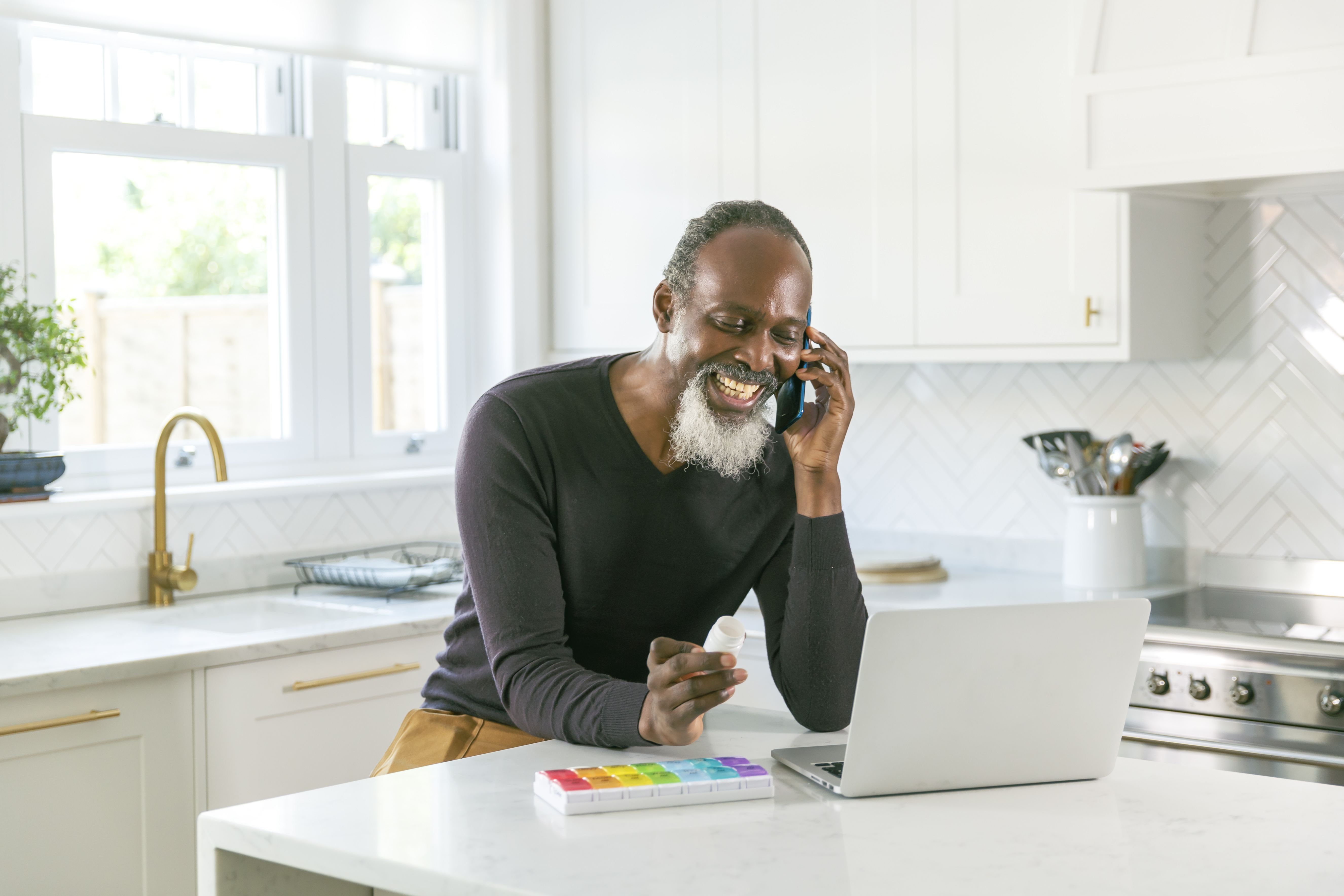 talking on phone_laptop_pill sorter_medication_senior male_diverse_iStock-1338748951